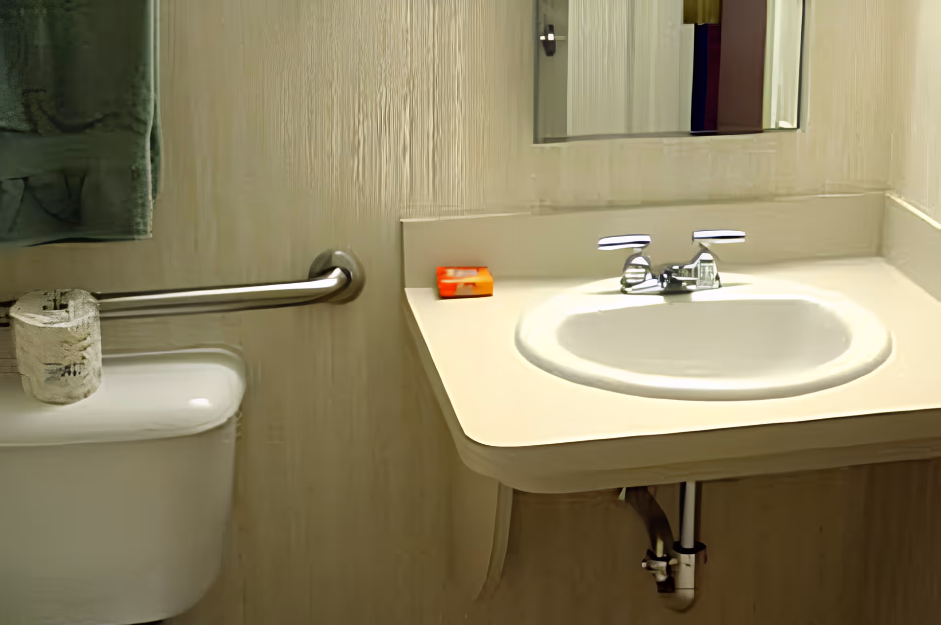 A small bathroom sink with a chrome faucet, a mirror above it, a bar of soap on the sink counter, a toilet with a roll of toilet paper on top, and a metal grab bar mounted on the wall next to the toilet.