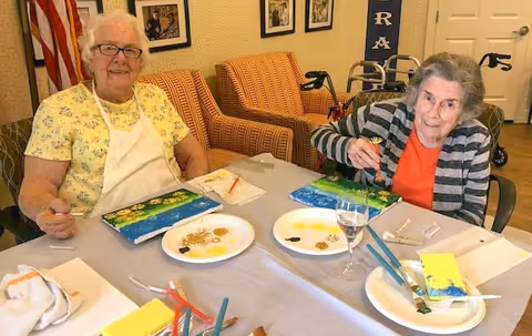 Two elderly women sitting at a table engaged in painting activities with brushes, paint palettes, and canvases in front of them. They are smiling and appear to be enjoying the activity in a cozy room with chairs and framed pictures on the wall.