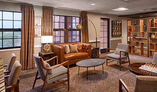 A cozy living room area with a brown leather sofa, two patterned armchairs, a round coffee table, and a side table with a lamp. The room has large windows with brown curtains, a bookshelf with decorative items, and a carpeted floor.