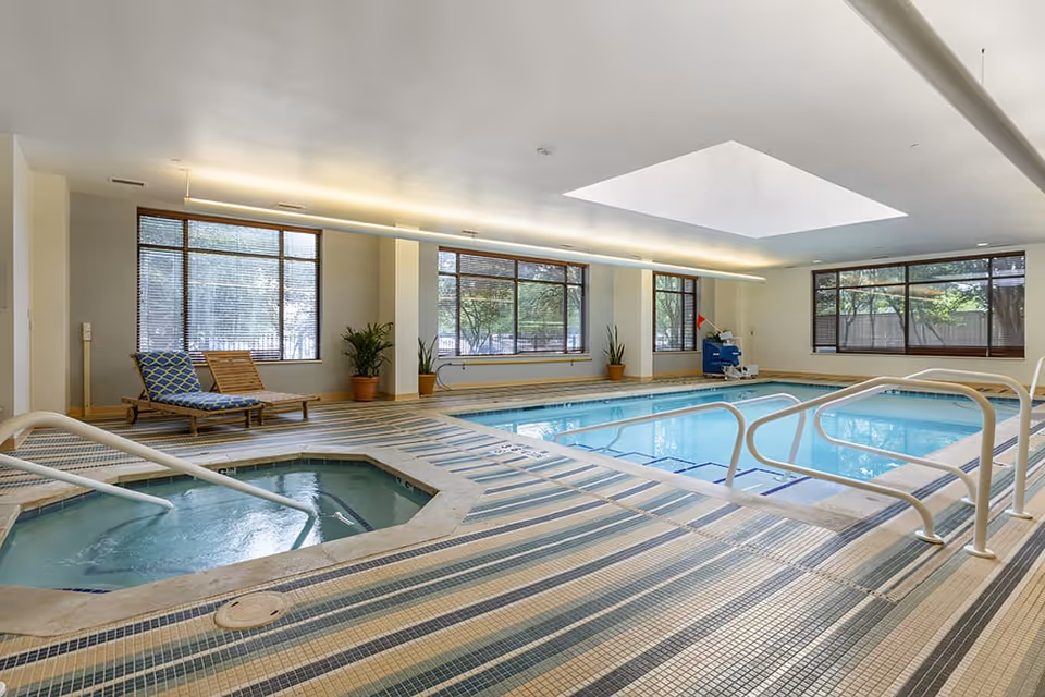 Indoor pool area with a rectangular swimming pool and a smaller hot tub. The floor is covered with striped tiles in shades of blue and beige. There are metal handrails for entering the pool and hot tub. Large windows with blinds let in natural light, and there are a couple of lounge chairs and potted plants along the wall. The ceiling has a large skylight and modern lighting fixtures.
