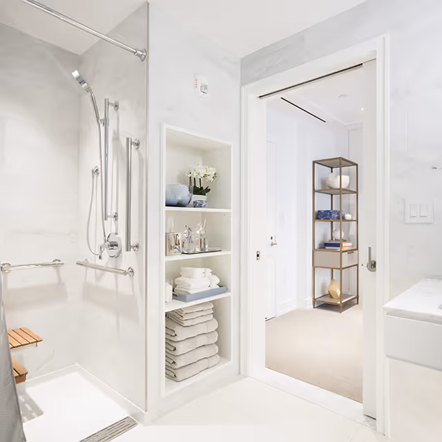 Bright and clean bathroom with a walk-in shower featuring a handheld showerhead and grab bars. Built-in shelves next to the shower hold neatly folded towels, decorative items, and toiletries. The bathroom opens into a carpeted room with a tall shelving unit displaying books and decorative objects.