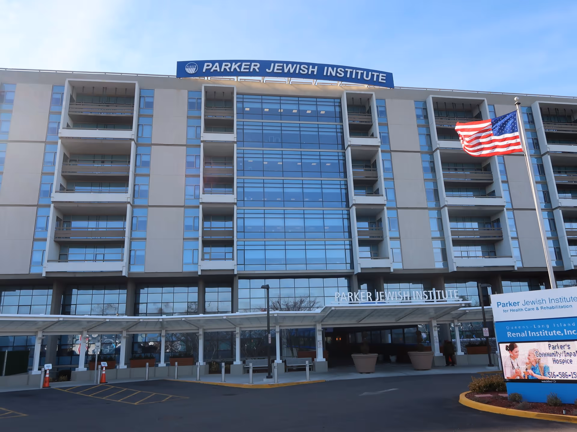 Exterior front view of the Parker Jewish Institute for Health Care & Rehabilitation building with multiple floors, large glass windows, balconies, an American flag on a pole, and a signboard displaying information about the facility.