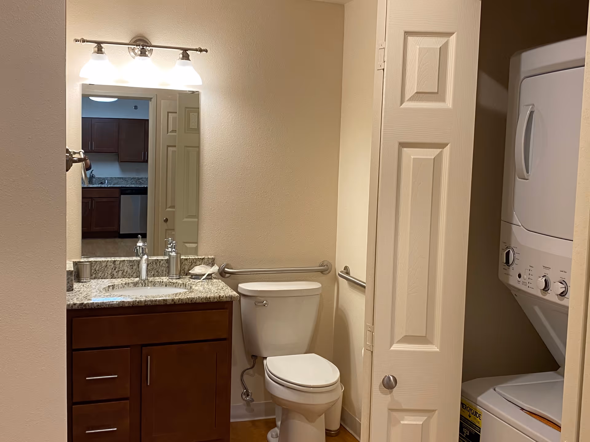 A small bathroom with a white toilet, a wooden vanity with a granite countertop and a sink, a mirror above the sink with three light fixtures, and a partially open door revealing a stacked washer and dryer unit.