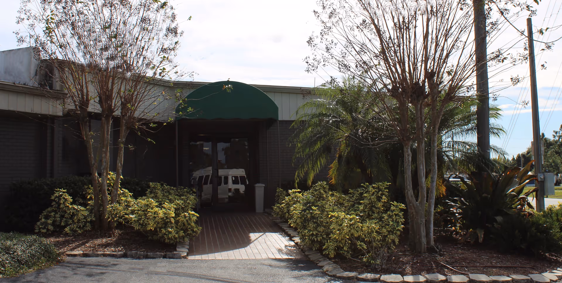 Entrance to a building with a green awning above the door, surrounded by bushes, small trees, and palm plants. A paved walkway leads to the glass double doors, and a white vehicle is reflected in the glass.