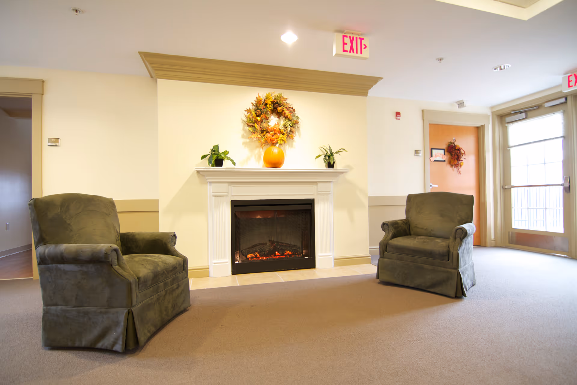 A cozy sitting area in a senior living facility with two green upholstered armchairs facing a white electric fireplace. Above the fireplace is a decorative autumn wreath and a pumpkin, with two small potted plants on either side. The room has beige walls, carpeted floor, and exit signs above the doors.