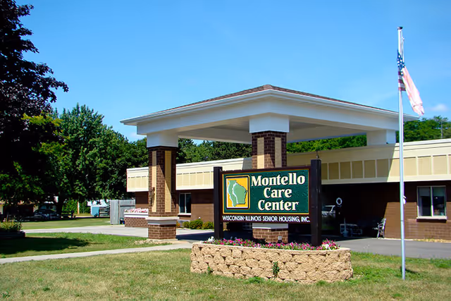 Exterior view of Montello Care Center building with a covered entrance supported by brick columns. A sign in front reads 'Montello Care Center Wisconsin-Illinois Senior Housing, Inc.' There is a flagpole with an American flag to the right and trees and grass surrounding the area under a clear blue sky.