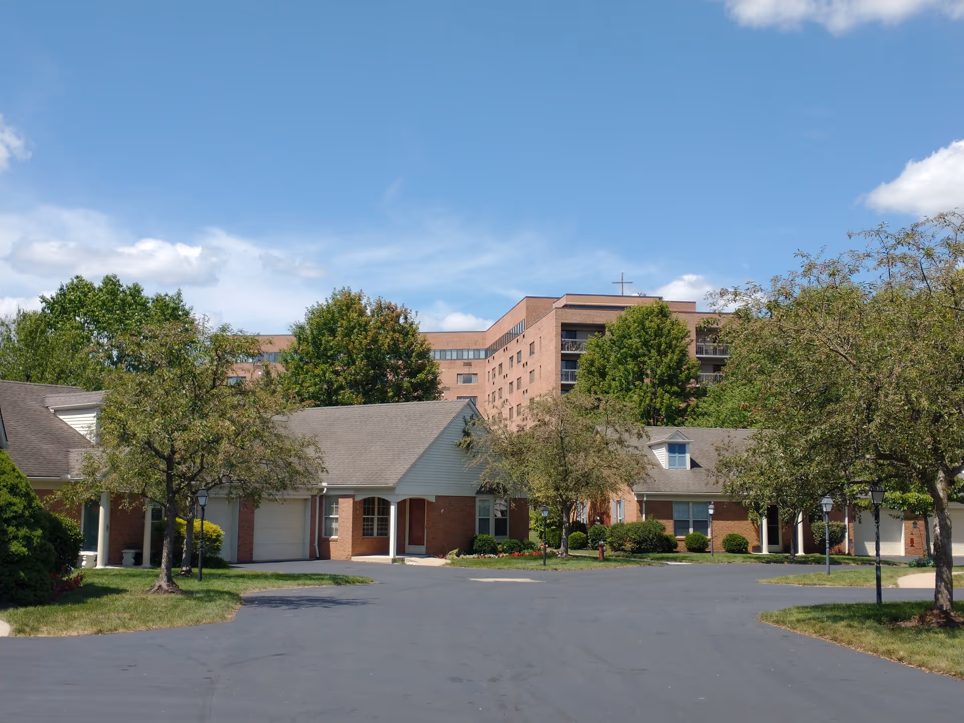 View of a senior living community with single-story brick cottages surrounded by trees and greenery, with a multi-story building in the background under a blue sky with some clouds.