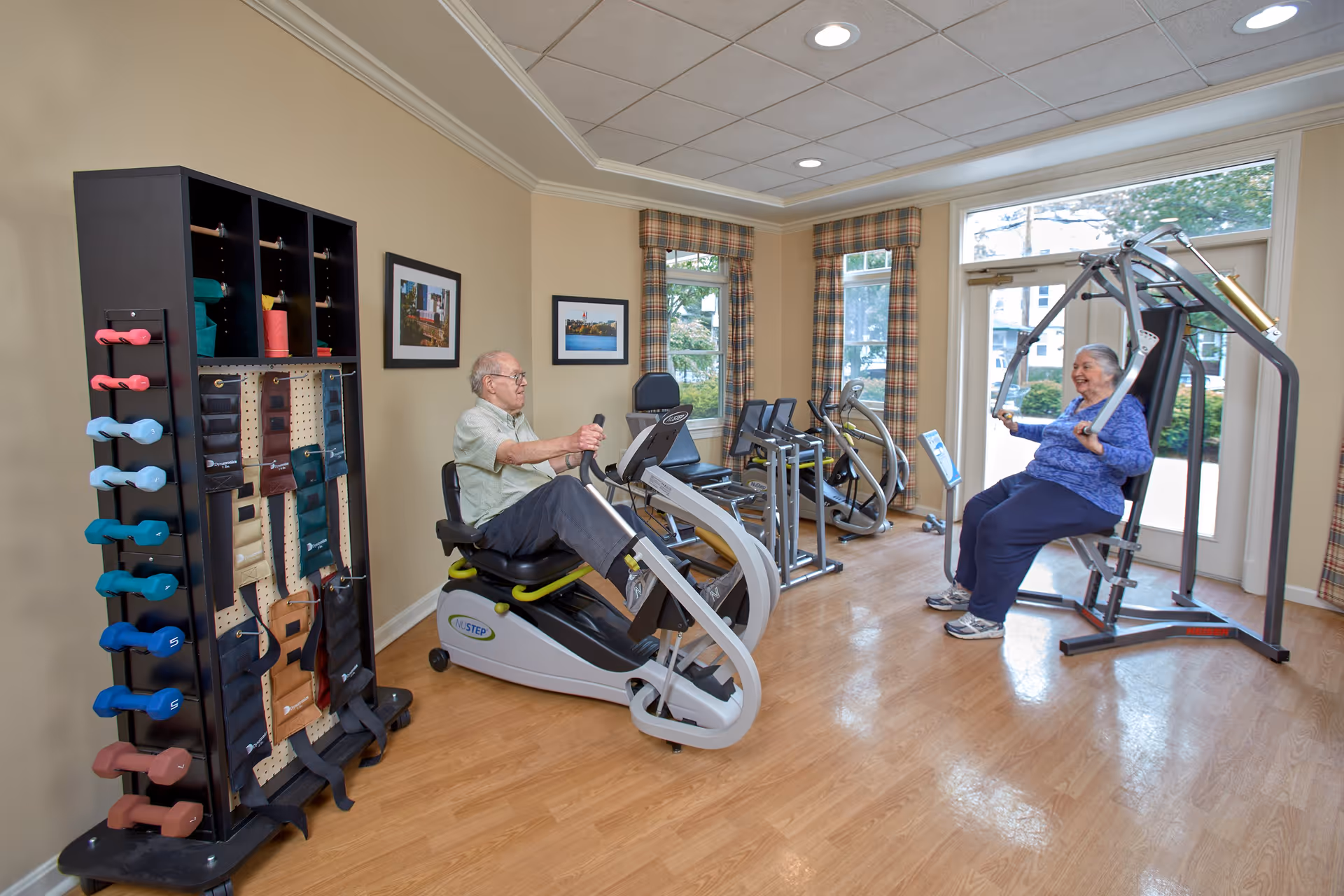 Two elderly individuals exercising in a well-lit fitness room with wooden floors. One man is using a recumbent exercise bike while a woman is seated on a strength training machine. The room has large windows with plaid curtains, framed pictures on the walls, and a rack holding various dumbbells and ankle weights.