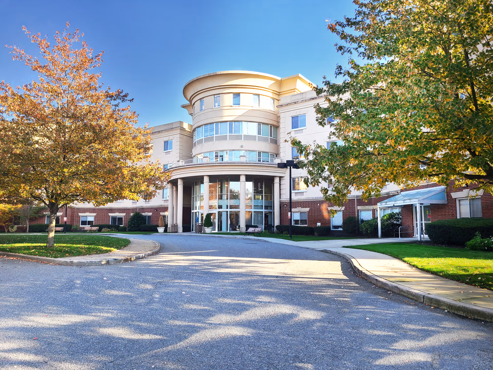 Front exterior view of Fulton Commons Care Center, a multi-story building with a rounded central entrance supported by columns. The building is surrounded by trees with autumn foliage and a paved driveway leading to the entrance.