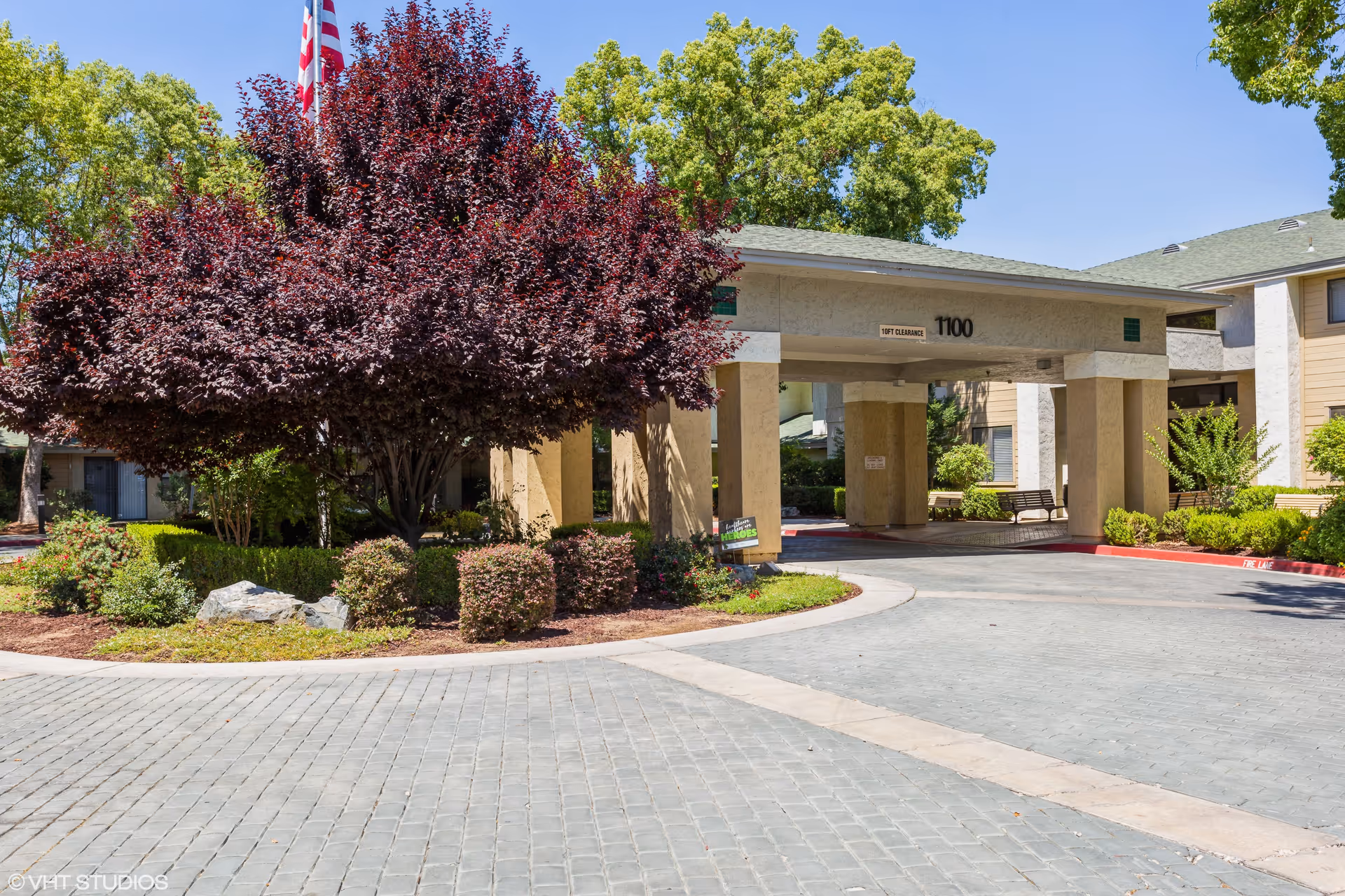 Entrance area of a senior living facility with a covered drop-off zone, surrounded by well-maintained landscaping including a large tree with dark red leaves, bushes, and green trees in the background under a clear blue sky.
