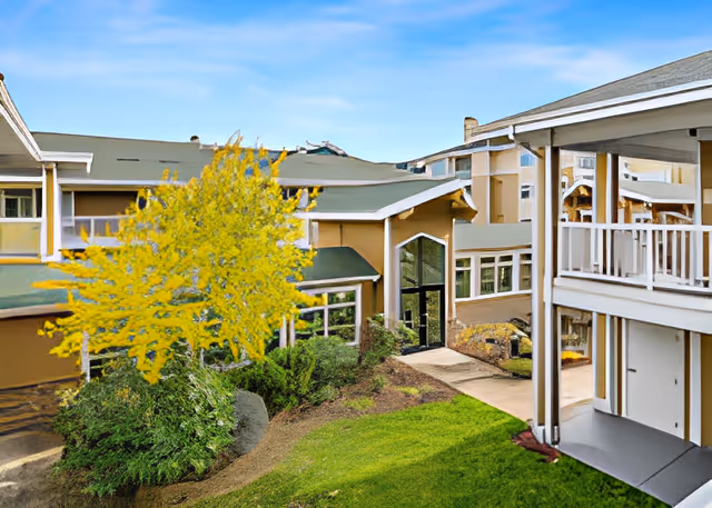 Exterior view of Cascade Manor senior living facility showing a courtyard with green grass, bushes, and a tree with yellow leaves. The building has beige walls, green roofs, and large windows. There is a covered balcony on the right side of the image.