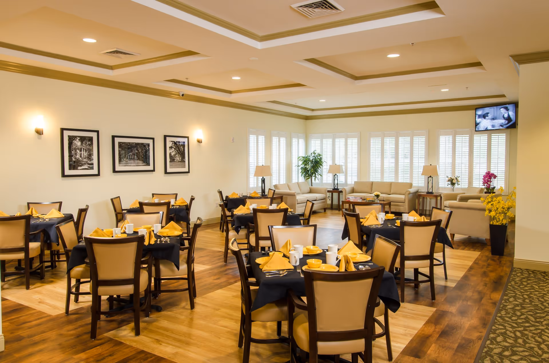 A bright dining room in a senior living facility with several tables set with yellow napkins, cups, and bowls. The room features wooden floors, beige walls with three black and white framed photos, and a seating area with beige sofas and lamps near large windows with white shutters. A small TV is mounted in the corner showing a black and white program.
