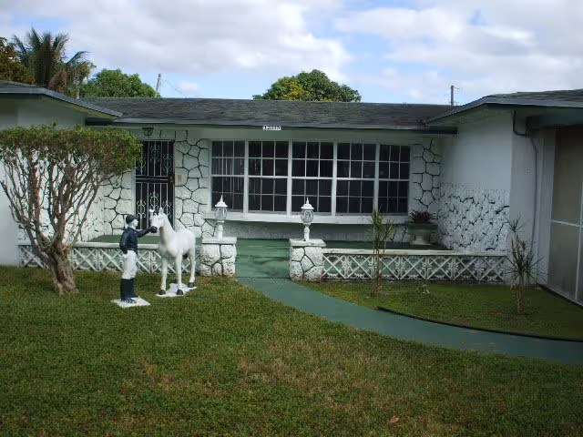 Front exterior view of a single-story building with white stone walls and a green pathway leading to the entrance. The lawn in front has two statues, one of a person and one of a horse, and there are small plants along the pathway.