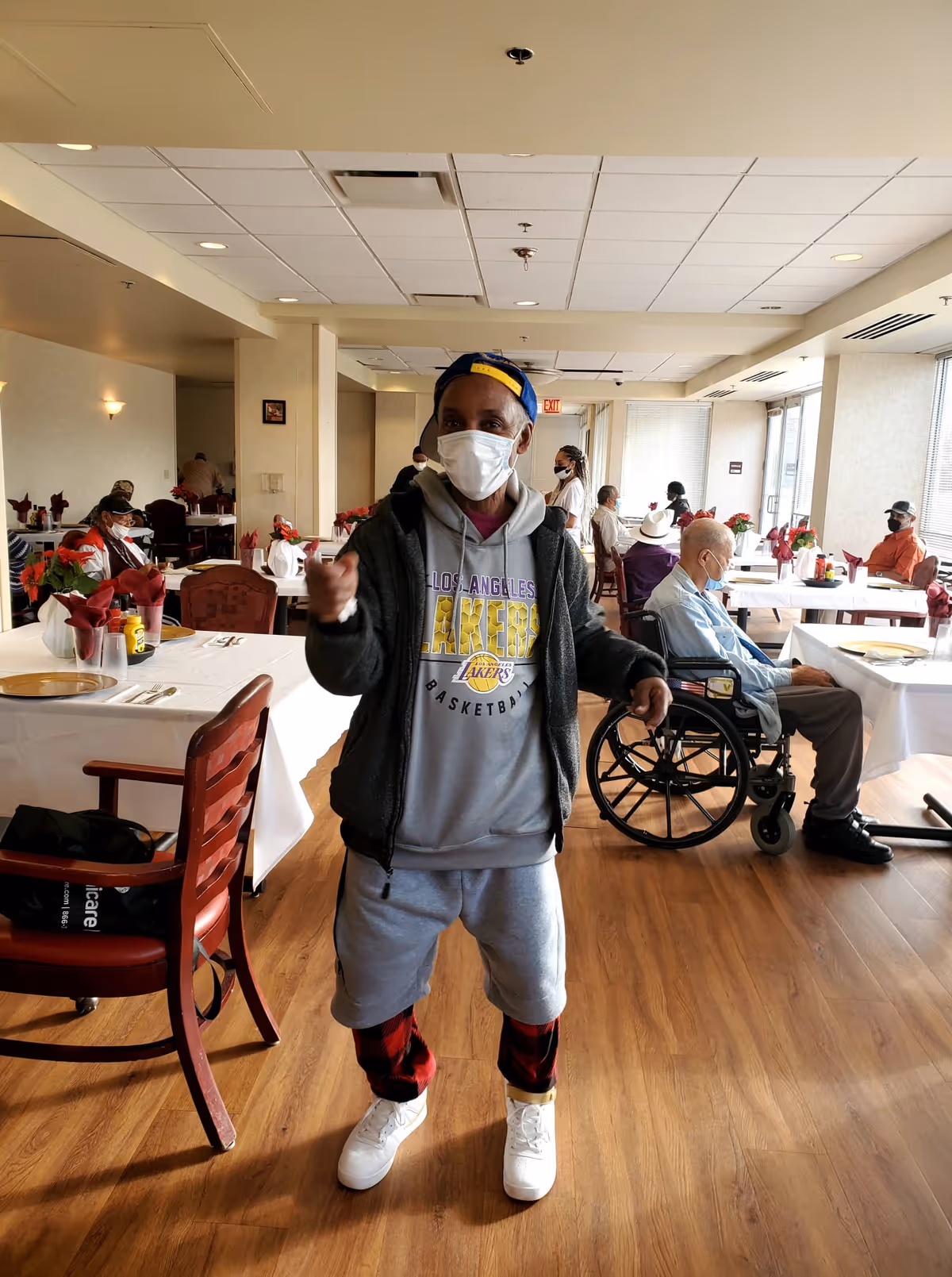 A person wearing a Los Angeles Lakers hoodie, gray shorts, and a face mask stands in a dining room with wooden floors. Several elderly people, some in wheelchairs, are seated at tables covered with white tablecloths and decorated with red napkins and flowers. The room has large windows letting in natural light.