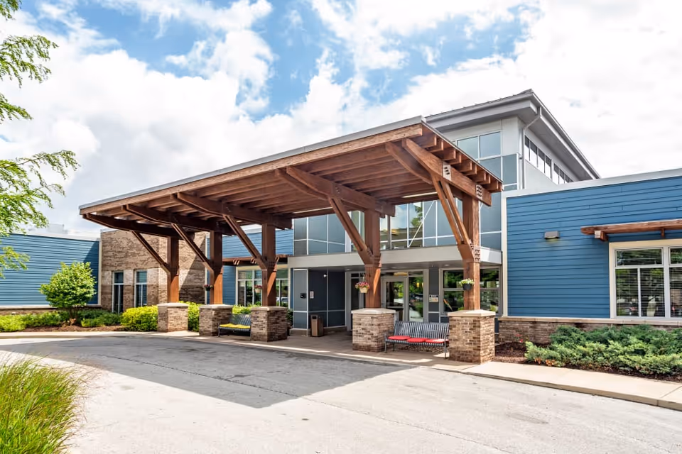Entrance of Arlington Place Health Campus featuring a large wooden canopy supported by brick pillars, blue siding on the building, large windows, benches under the canopy, and landscaped greenery around the building under a partly cloudy sky.
