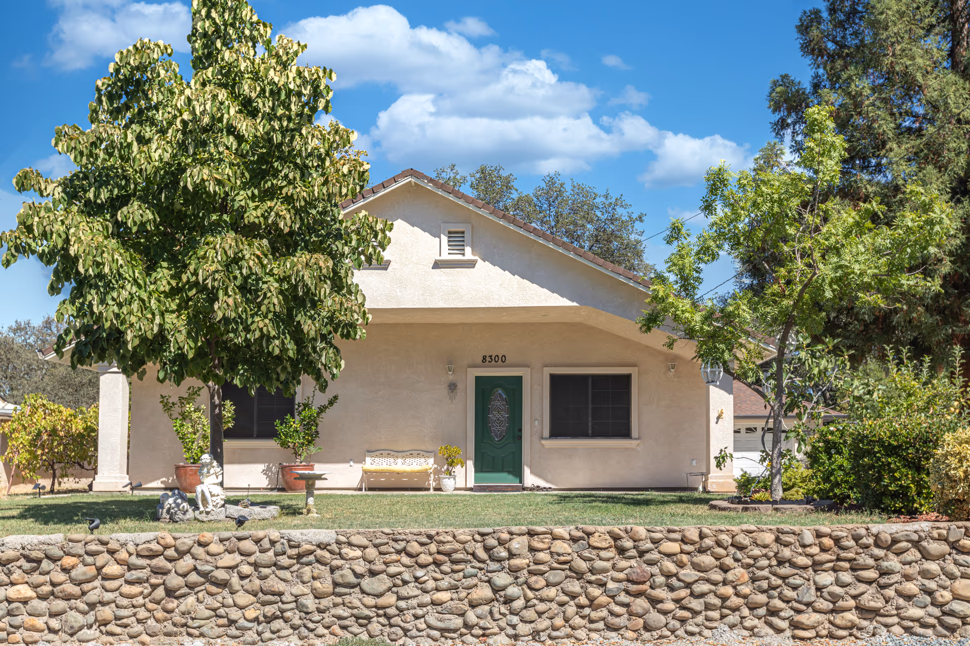 Front exterior view of a single-story house with a green door, two windows, a stone wall in the foreground, and trees on either side under a blue sky with clouds.