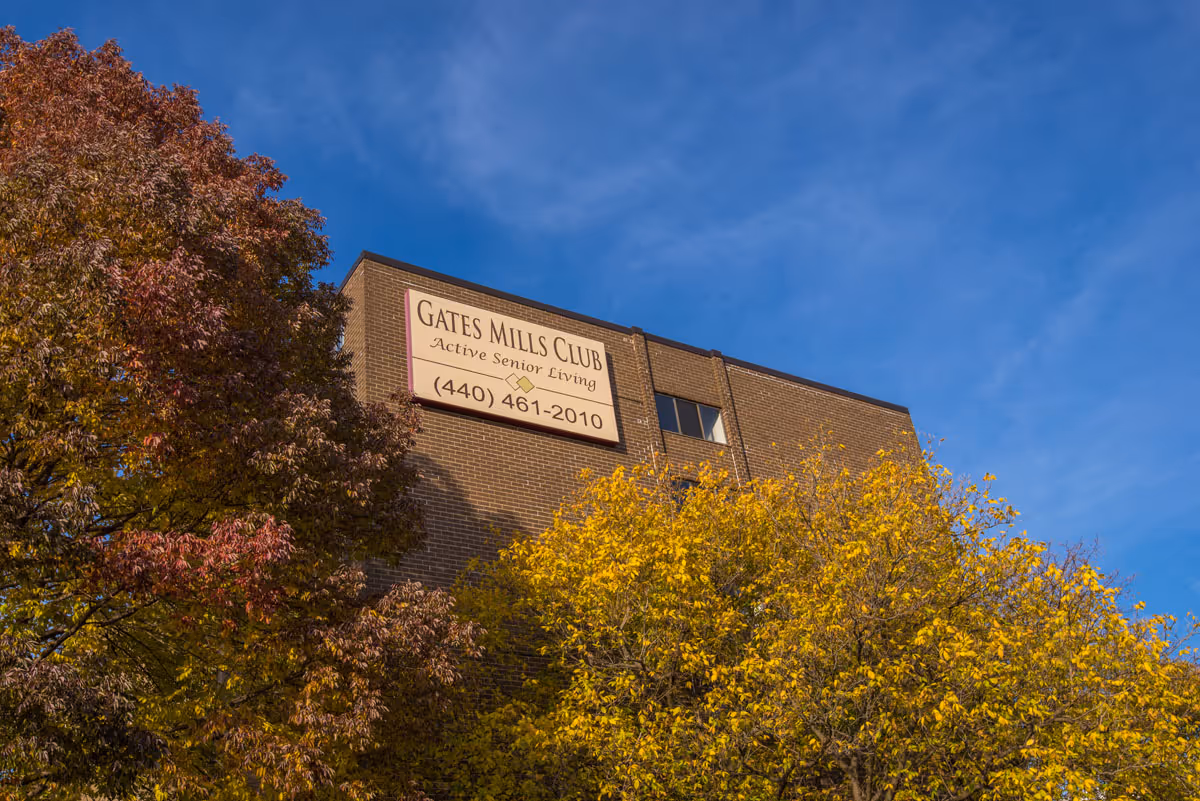 Exterior view of a brick building with a sign that reads 'Gates Mills Club Active Senior Living (440) 461-2010' partially obscured by trees with autumn foliage under a clear blue sky.