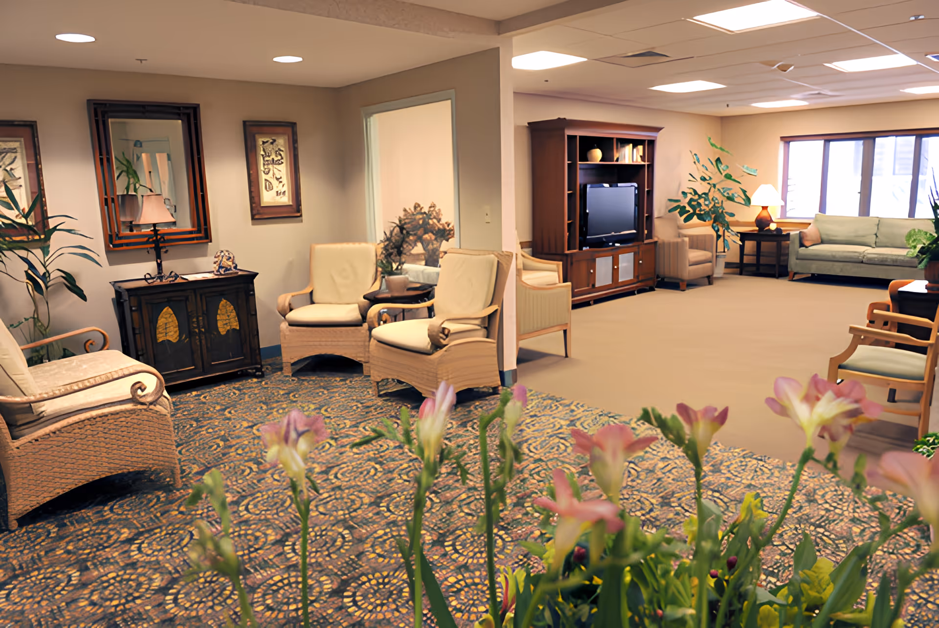 A cozy living room area in Springfield Crossings featuring cushioned wicker chairs, a patterned carpet, a wooden entertainment center with a TV, a sofa, armchairs, side tables with lamps, framed artwork on the walls, and potted plants. Flowers are visible in the foreground.