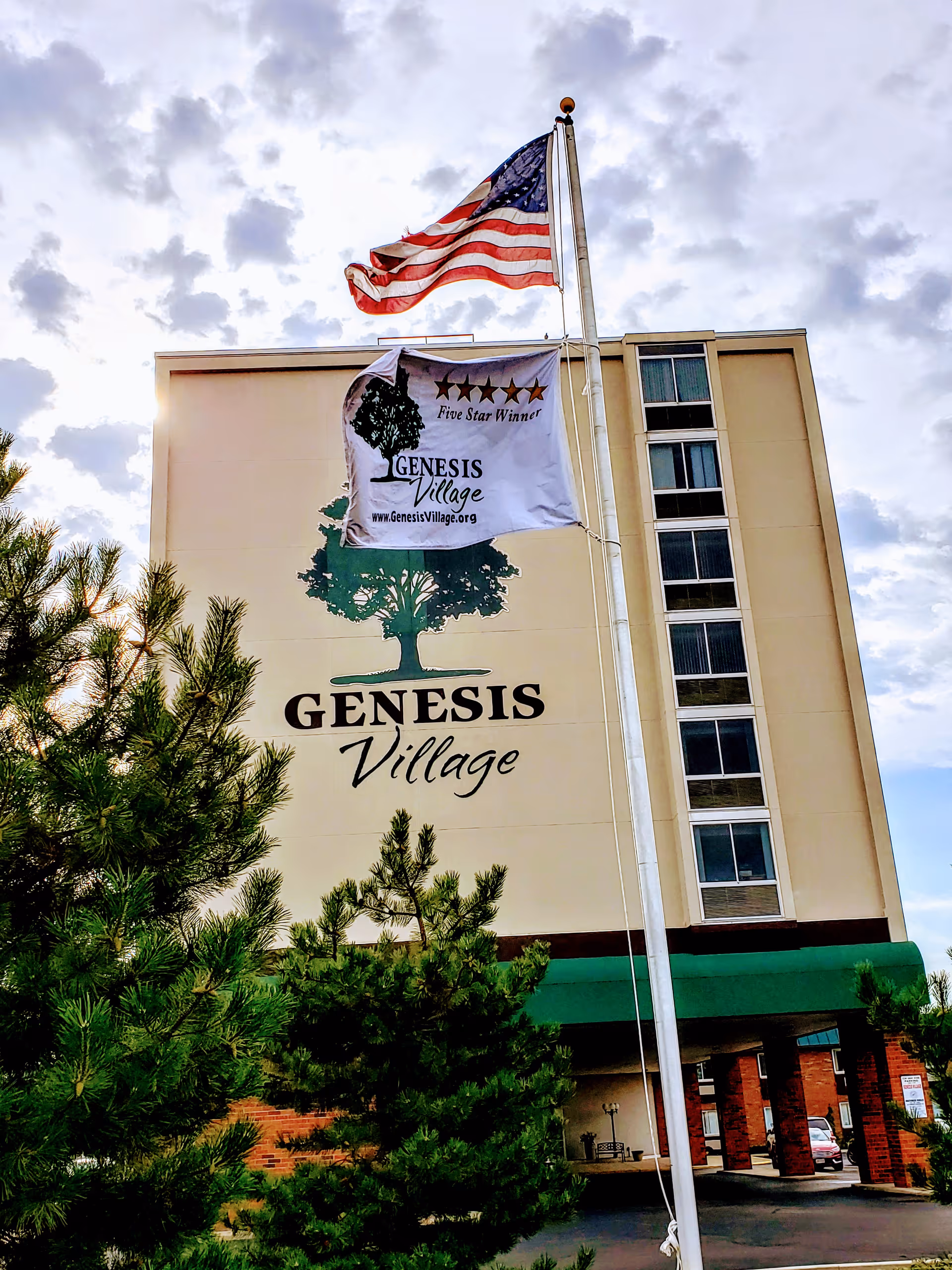 Exterior view of the Genesis Village building with a large tree logo and the name displayed on the wall. An American flag and a Genesis Village flag with a five-star winner emblem are flying on a flagpole in front of the building. Green trees are visible in the foreground under a cloudy sky.
