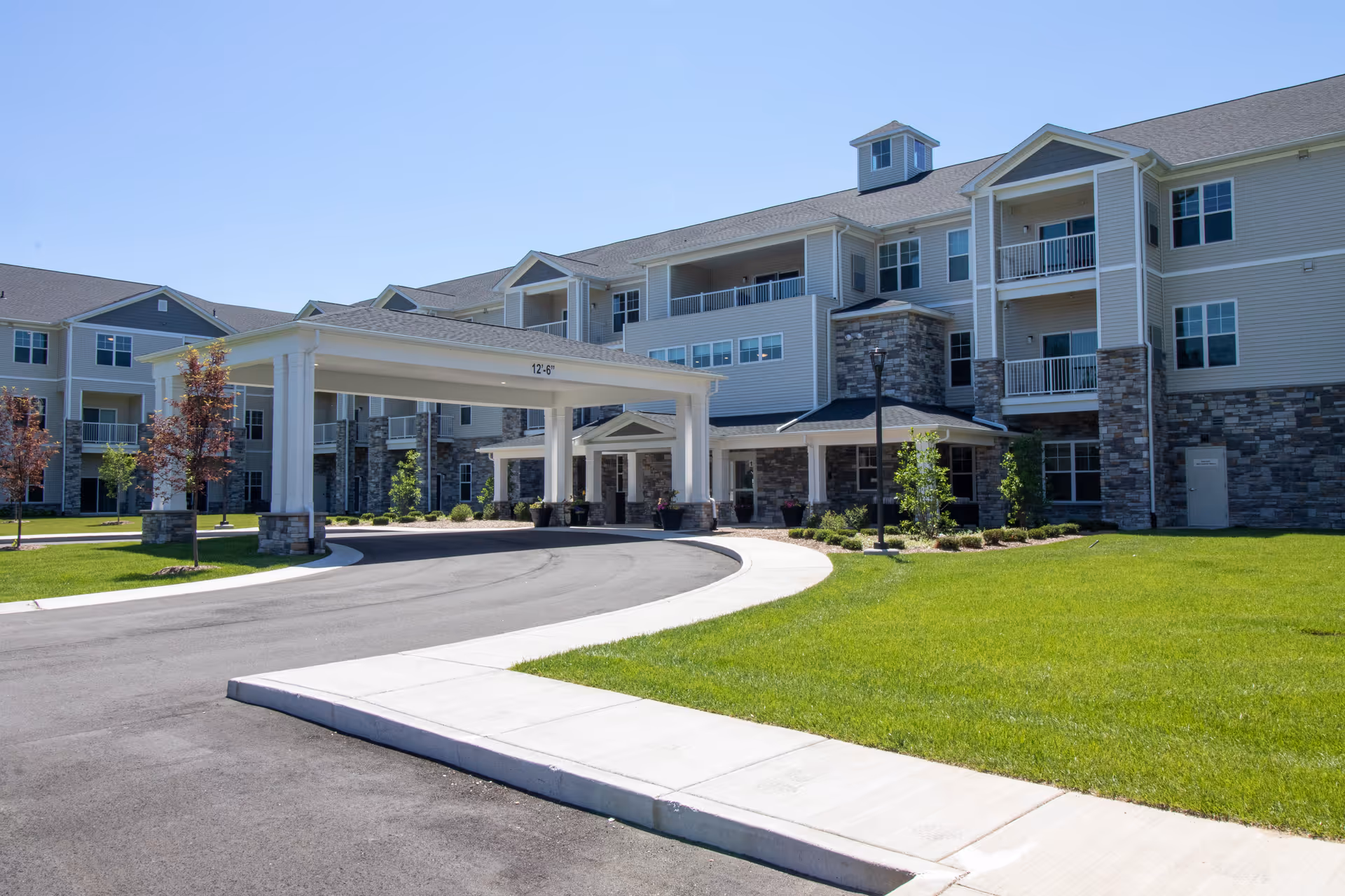 Exterior view of a large, modern senior living facility building with multiple floors, balconies, and a covered entrance driveway. The building features stone and siding facade with well-maintained green lawns and small trees under a clear blue sky.