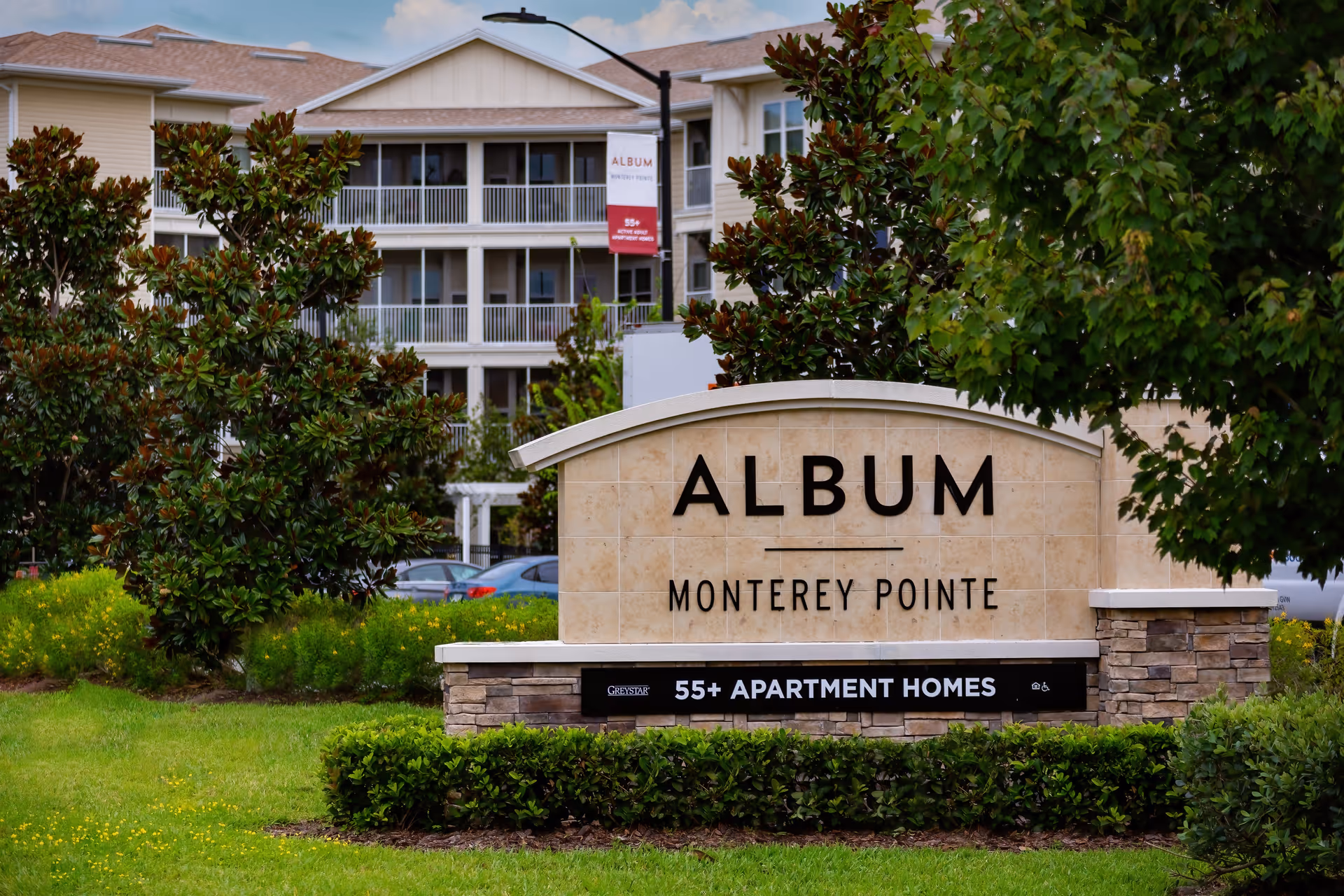 Outdoor view of the entrance sign for Album Monterey Pointe, a 55+ apartment home community, with a multi-story residential building and greenery in the background.