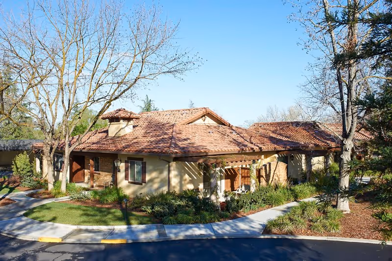 Exterior view of a single-story building with a tiled roof and beige walls, surrounded by trees and landscaped greenery under a clear blue sky.