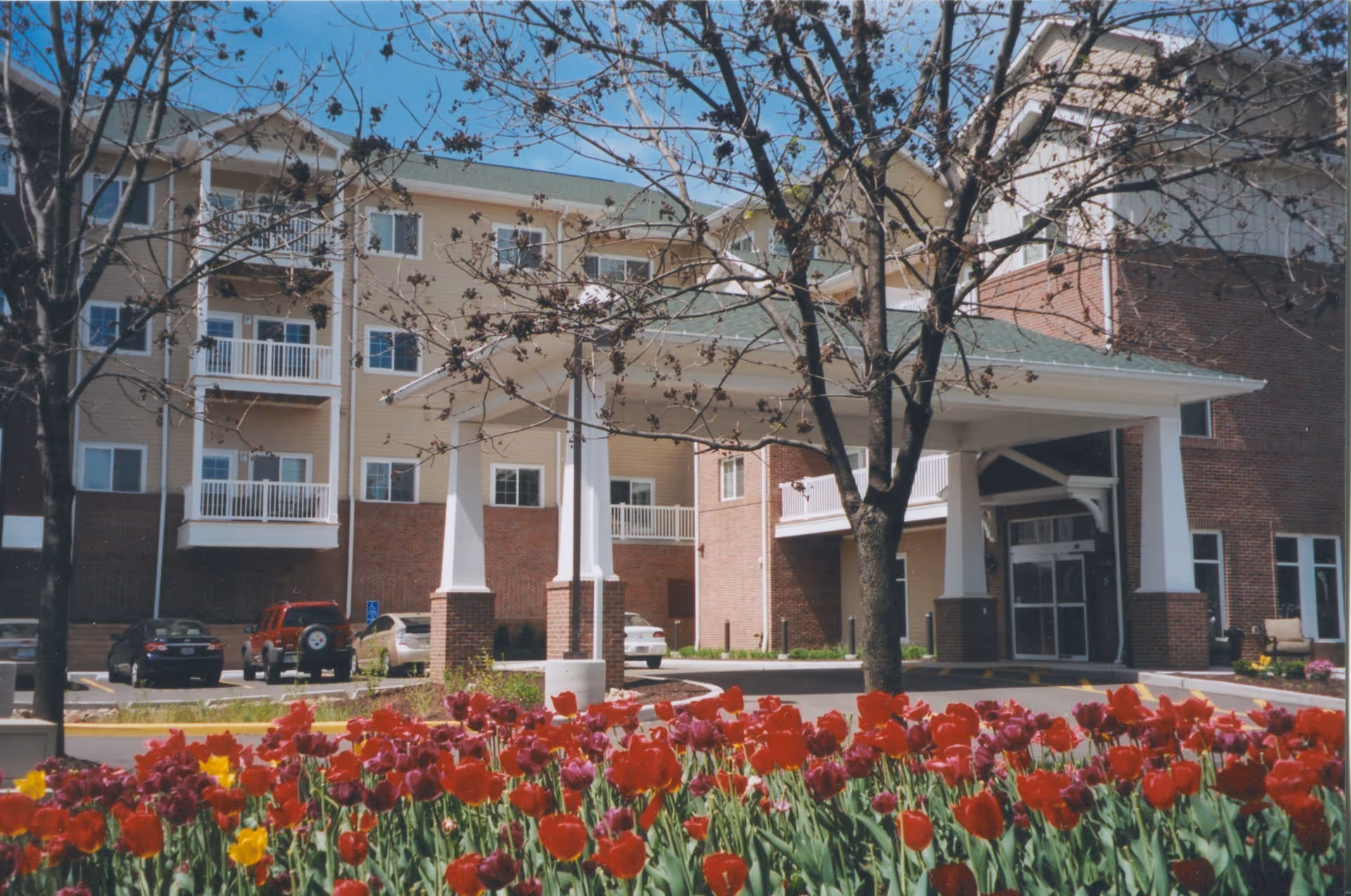 Exterior view of Nazareth Living Center showing a multi-story building with balconies, a covered entrance supported by white columns, several parked cars, leafless trees, and a vibrant flower bed with red, purple, and yellow tulips in the foreground under a clear blue sky.
