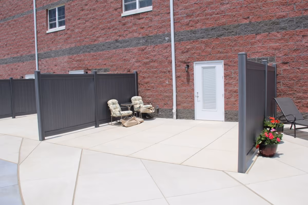 Outdoor patio area with concrete flooring, two cushioned chairs and a small table against a red brick wall with a white door. There are privacy panels and potted flowers near a lounge chair.