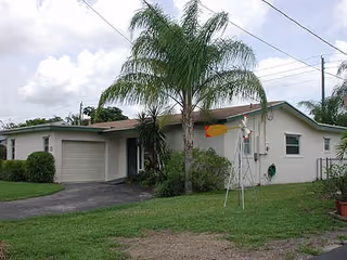 Single-story residential building with a light-colored exterior, a garage on the left side, a palm tree and other greenery in front, and a small windmill decoration on the lawn under a partly cloudy sky.