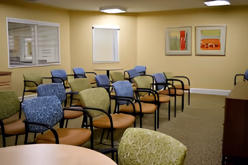 A room with multiple rows of chairs arranged facing forward, featuring a mix of blue and green upholstered seats with black armrests. The walls are beige with two abstract paintings hanging on one side and a window with a closed blind. There is a round table with a green chair in the foreground and a wooden cabinet on the right side.