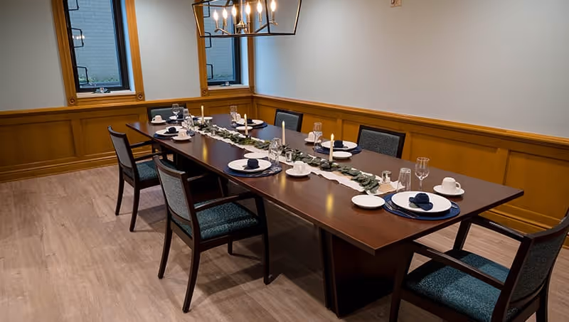 A formal dining room with a long wooden table set for six people. The table is decorated with a white runner and greenery centerpiece, and each place setting includes a plate, napkin, cup, and glass. The room has wood paneling on the walls, two windows, and a modern chandelier hanging above the table.