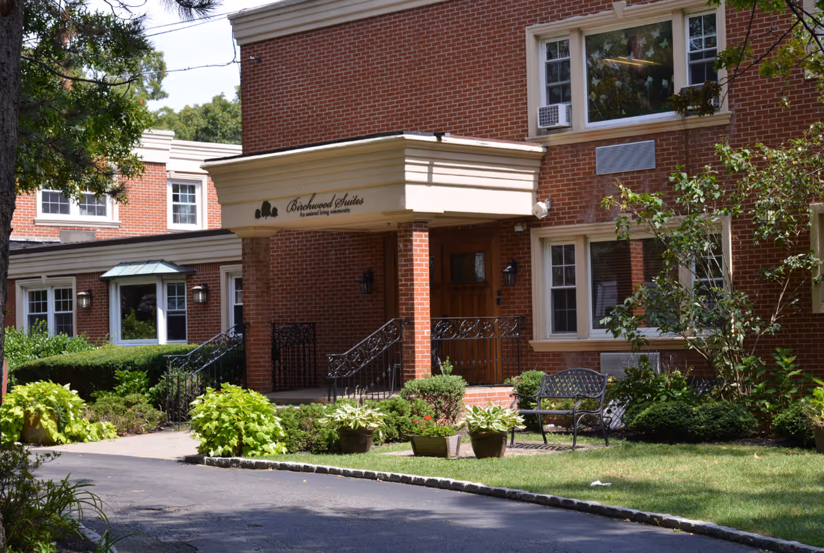 Exterior view of Birchwood Suites, an assisted living community, showing a brick building entrance with a covered porch, black wrought iron railings, and surrounding greenery including bushes and potted plants. A black metal bench is placed on the lawn near the entrance.