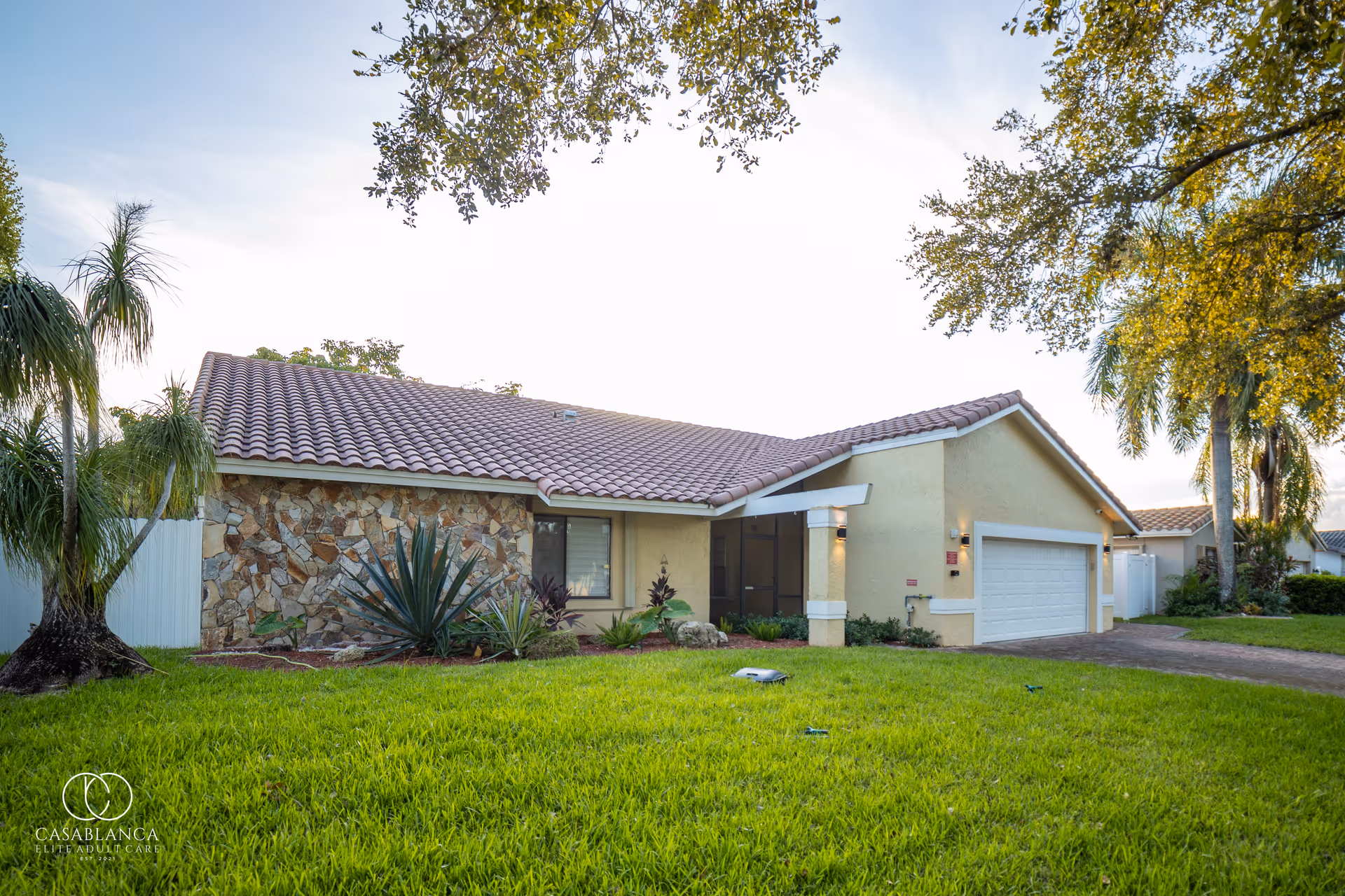 Exterior view of a single-story residential building with a tiled roof, stone and stucco walls, a two-car garage, and a well-maintained green lawn with various plants and trees around.