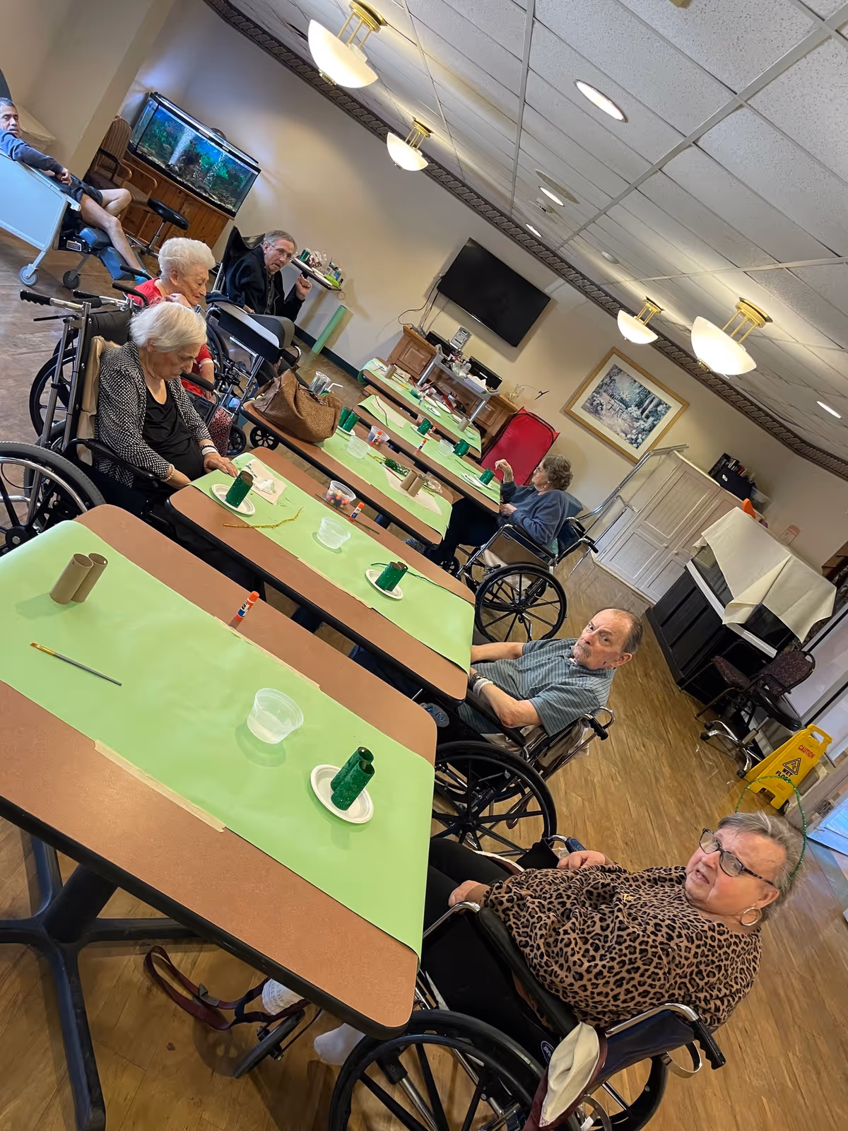 A group of elderly people sitting in wheelchairs around tables covered with green paper in a well-lit room. The room has a wooden floor, a large TV mounted on the wall, a fish tank, and various furniture including a piano and chairs. The elderly individuals appear to be engaged in an activity or social gathering.