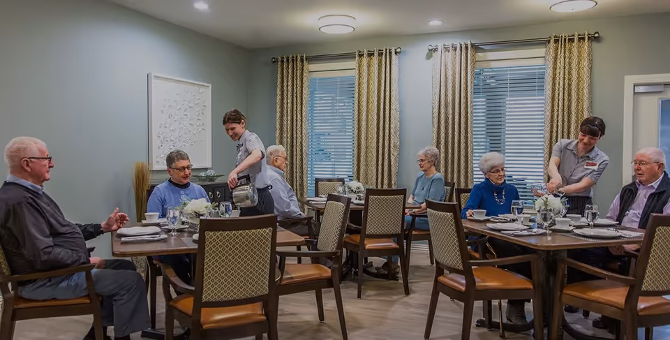 A dining room in a senior living facility with elderly residents seated at tables. Two staff members are serving drinks to the residents. The room has light-colored walls, patterned curtains, and wooden chairs with cushioned seats.
