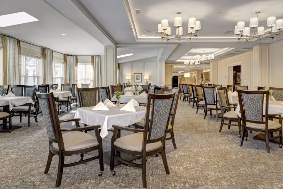 A spacious, well-lit dining room with multiple tables covered in white tablecloths, each set with folded napkins and small flower arrangements. The room features patterned chairs with wooden frames, large windows with curtains allowing natural light, and elegant ceiling light fixtures.