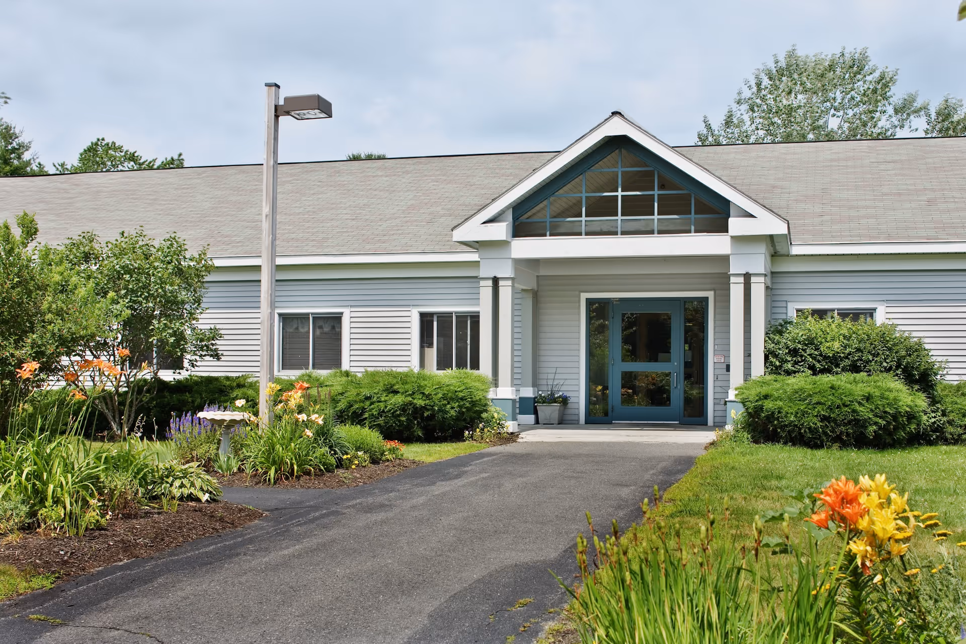 Front exterior view of Pine Point Center, a single-story building with light gray siding and a peaked roof. The entrance features double glass doors under a small portico with white columns. The walkway leading to the entrance is surrounded by well-maintained green shrubs, flowering plants, and a lamp post on the left side.