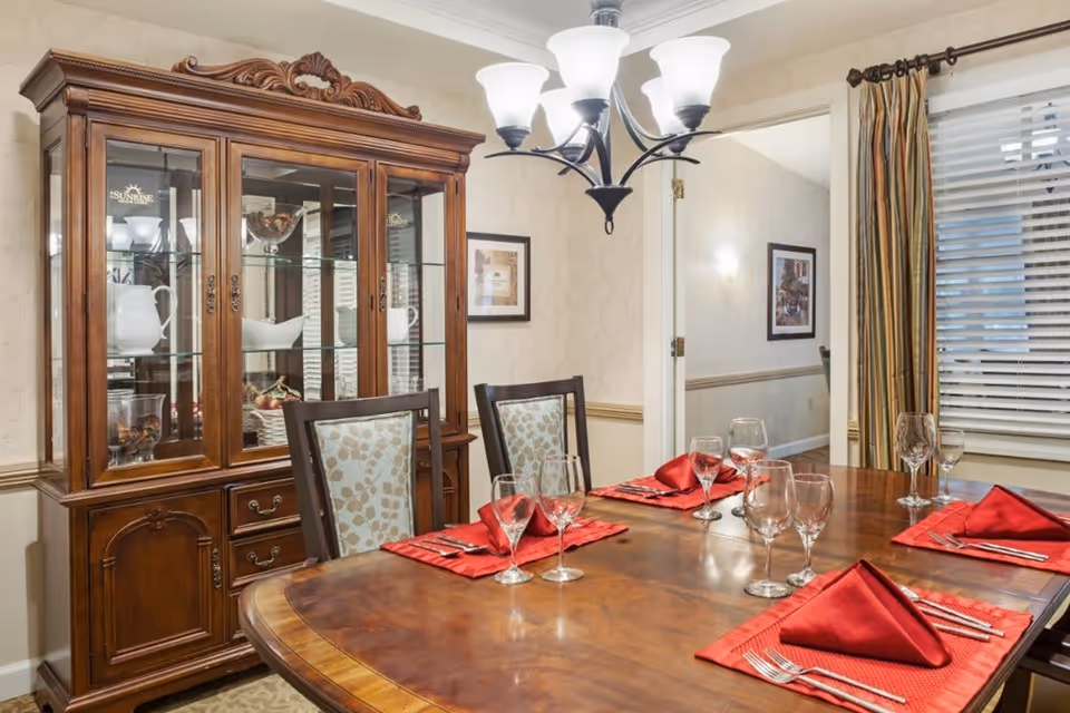 A formal dining room with a wooden dining table set for six, featuring red napkins, glassware, and silverware. Behind the table is a wooden china cabinet displaying white dishes and decorative items. The room has beige walls, a chandelier with five lights, a window with striped curtains, and framed artwork on the walls.