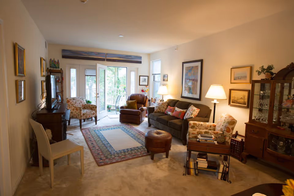 A cozy living room with a brown sofa adorned with colorful pillows, two patterned armchairs, a brown recliner, and a round leather ottoman. The room features a patterned area rug, a wooden cabinet with glassware, framed artwork on the walls, a TV on a wooden stand, and a door leading to a balcony with outdoor seating. The space is warmly lit by table lamps and natural light from windows.