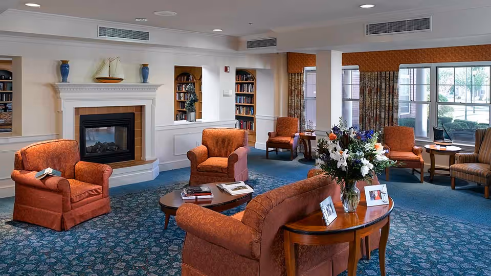 A cozy living room area in a senior living facility with orange upholstered armchairs and a sofa arranged around a wooden coffee table and a side table with a vase of flowers. There is a fireplace with decorative items on the mantel, built-in bookshelves, large windows with patterned curtains, and a blue patterned carpet.