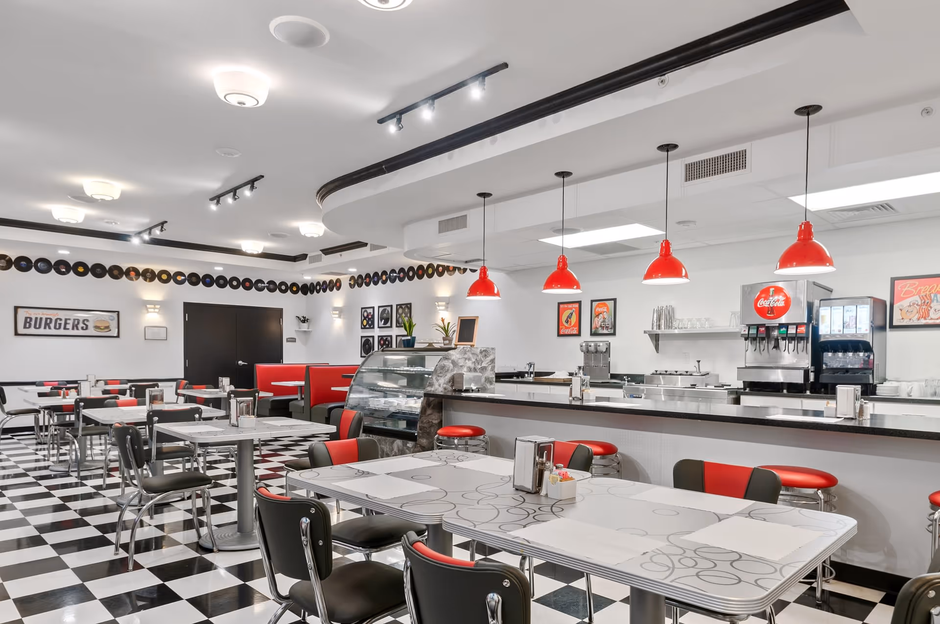 Interior of a retro-style diner with black and white checkered floor, red and black chairs, red booth seating, and a counter with red pendant lights. The walls are decorated with vintage posters and a row of vinyl records near the ceiling. A soda fountain and condiment dispensers are visible behind the counter.