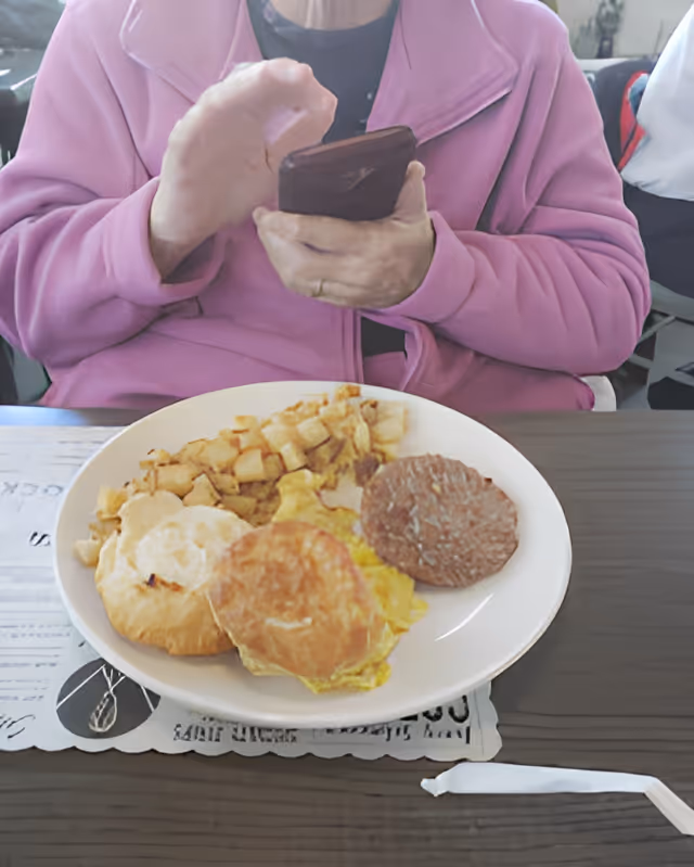 A person wearing a pink jacket is holding a smartphone above a plate of breakfast food, which includes scrambled eggs, a sausage patty, diced potatoes, and two biscuits, on a table with a placemat and a wrapped straw.