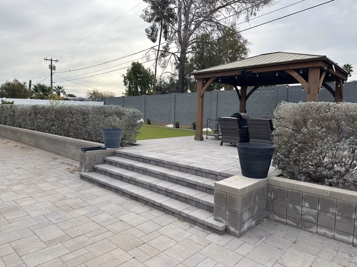 Outdoor patio area with a wooden pergola covering wicker chairs and a table. The patio is elevated with a set of stone steps leading up to it, surrounded by low concrete block walls and shrubbery. The sky is overcast and there are power lines and trees in the background.
