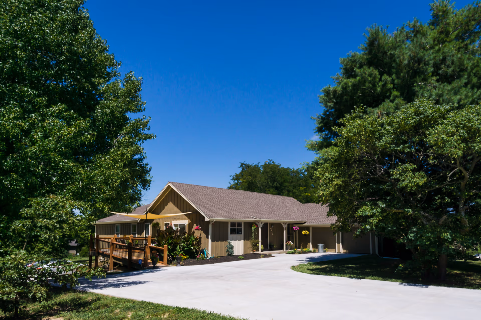 Single-story residential building with a sloped roof surrounded by trees and greenery under a clear blue sky. There is a concrete driveway leading up to the building and a wooden ramp on the left side with plants and hanging flower pots near the entrance.