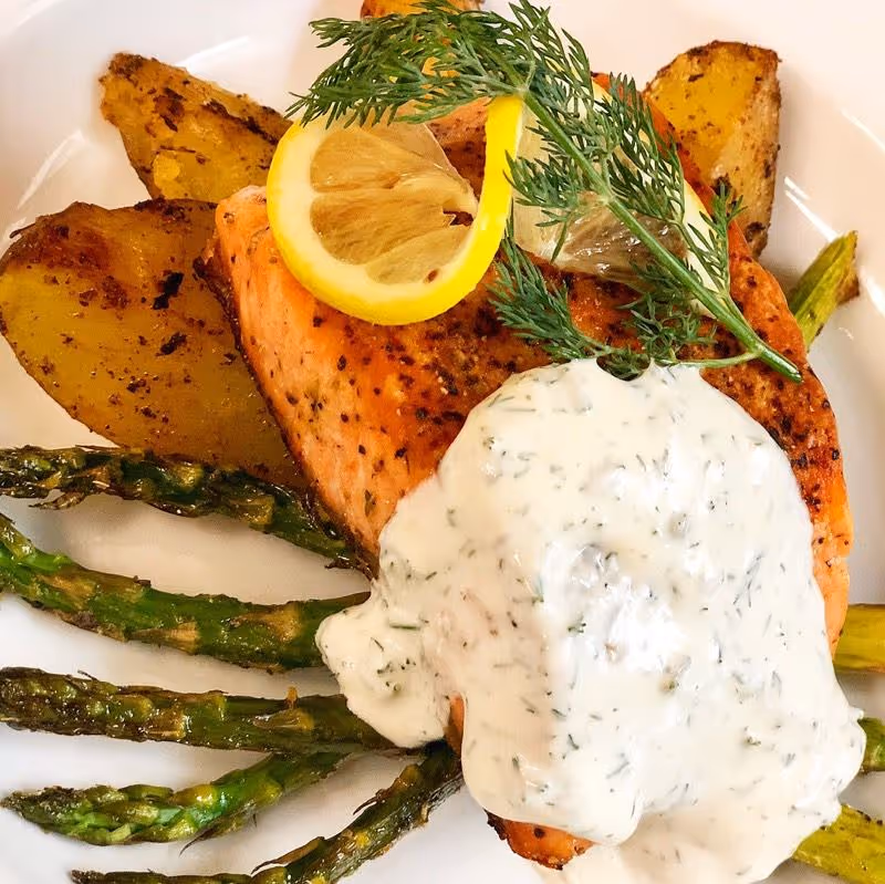 A plated meal featuring a seasoned salmon fillet topped with a lemon slice and fresh dill, accompanied by roasted potato wedges and grilled asparagus, with a creamy herb sauce drizzled over the salmon.