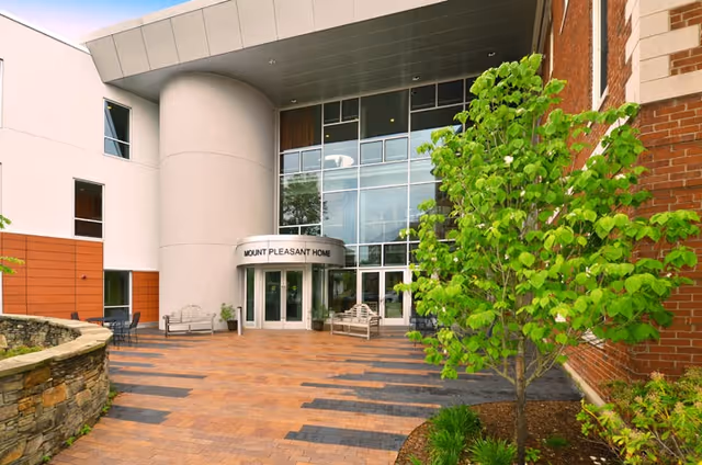 Entrance of Mount Pleasant Home featuring a modern building facade with large glass windows, a curved canopy above the double glass doors, outdoor benches, a small tree, and a stone wall planter on a paved courtyard.