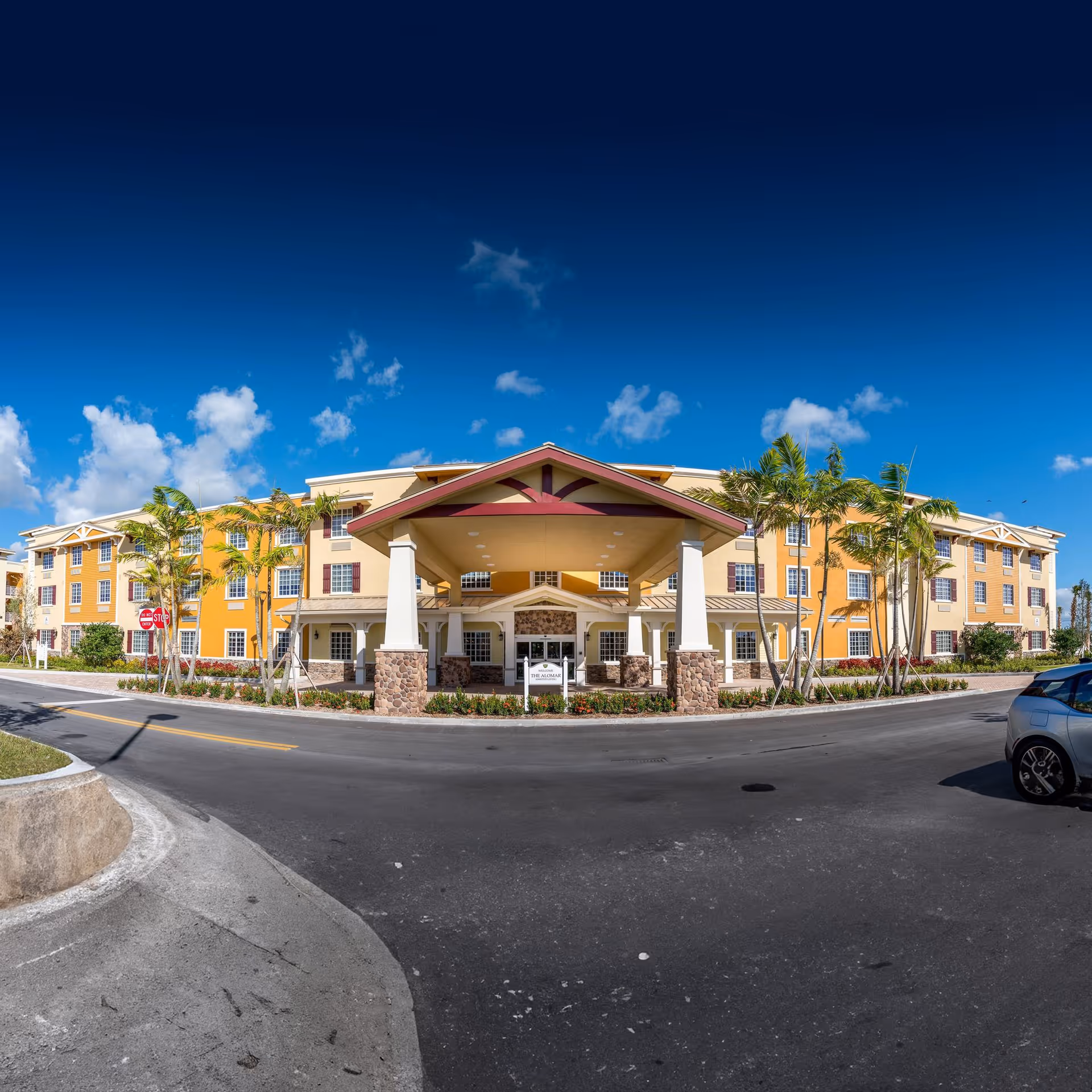 Front exterior view of Oakmonte Village of Davie, a large three-story building with a covered entrance supported by stone and white pillars. The building is painted in yellow and beige tones with red window shutters. Palm trees and landscaping surround the entrance area under a clear blue sky.
