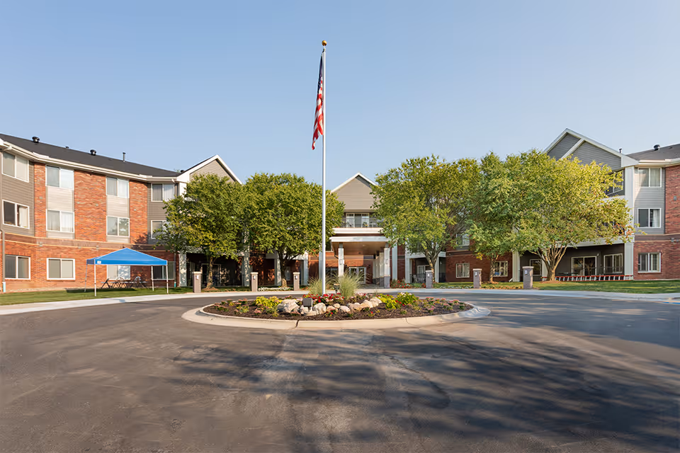Front exterior view of Immanuel: Arboretum Village, a multi-story senior living facility with a circular driveway, landscaped roundabout with flowers and rocks, several trees, and an American flag on a flagpole in the center.