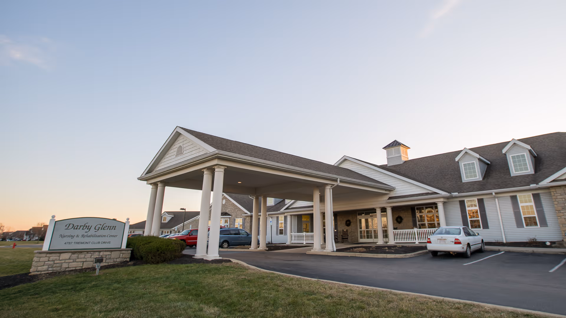 Exterior view of Darby Glenn Nursing & Rehabilitation Center at sunset, showing a large covered entrance with white columns, a parking area with several cars, and a sign with the facility's name and address on a grassy lawn.