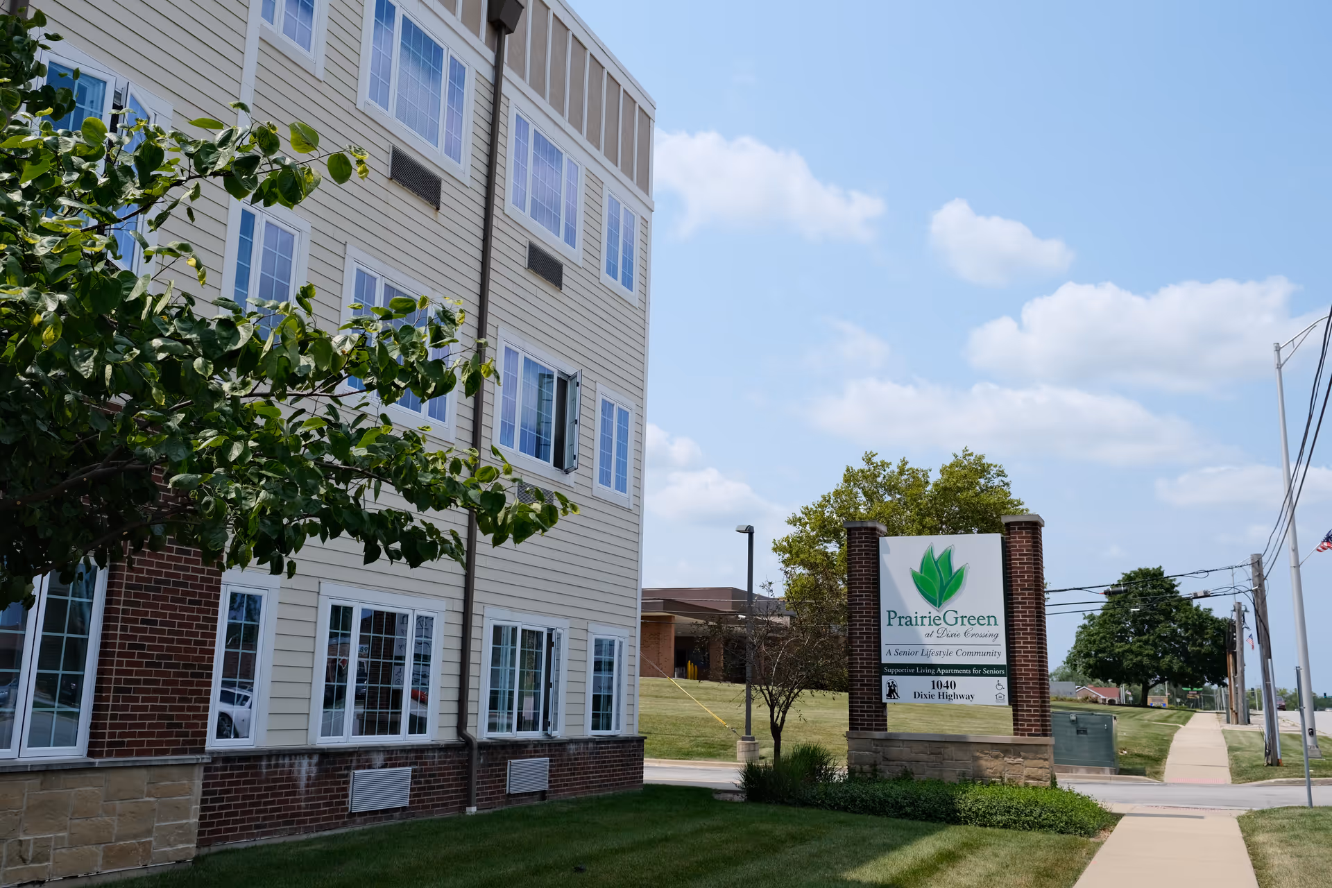 Exterior view of a senior living facility building with multiple windows, a tree in the foreground, and a sign that reads 'Prairie Green at Dixie Crossing, A Senior Lifestyle Community, Supportive Living Apartments for Seniors, 1040 Dixie Highway'. The sky is partly cloudy.
